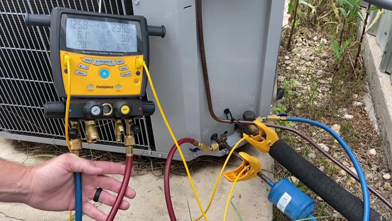 Digital manifold gauge connected to an outdoor AC unit, with blue and red hoses attached to the service ports, yellow temperature clamps on the suction and liquid lines, and the Fieldpiece display showing live readings.