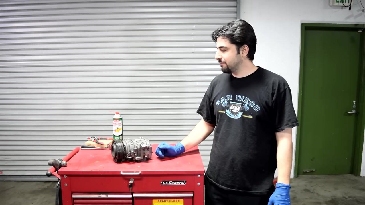 A person wearing blue gloves stands next to a red tool chest with an AC compressor, a can of CRC Brakleen, and various hand tools on top. The workspace is a garage with a closed roll-up door and a green exit door in the background.