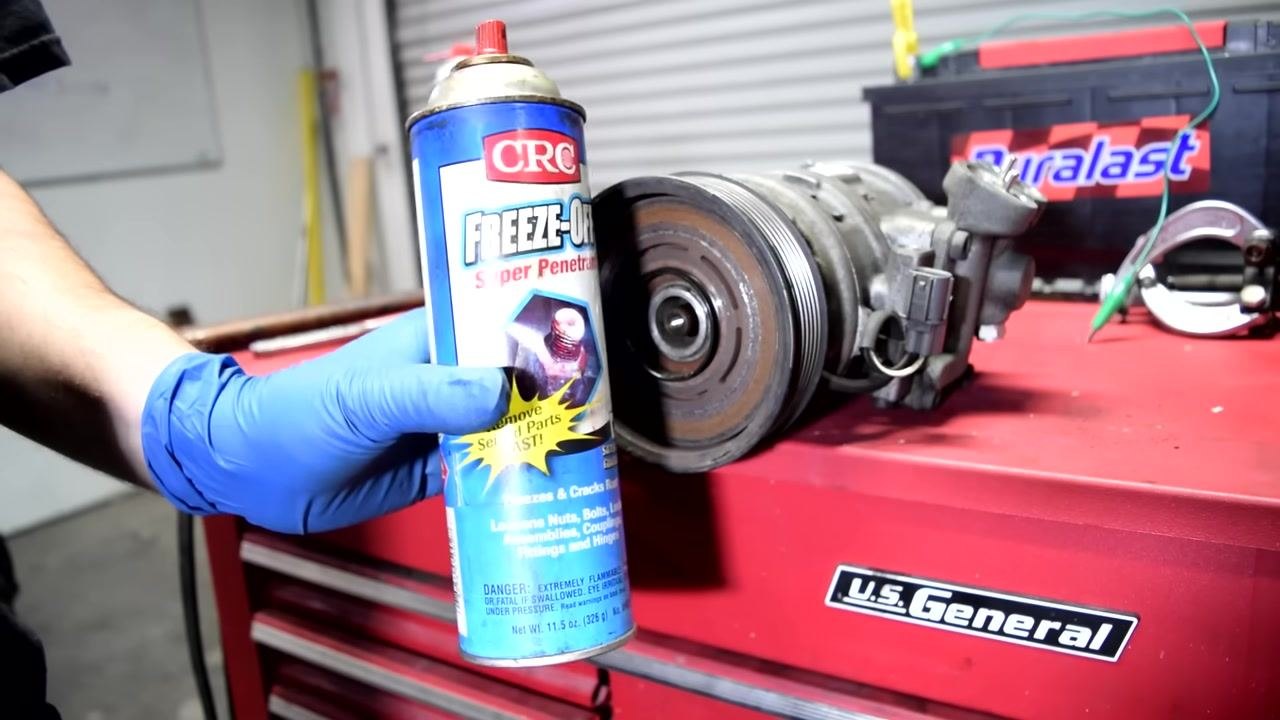 A person in blue gloves holds a can of CRC Freeze-Off Super Penetrant next to the AC compressor and pulley, preparing to spray the fluid.