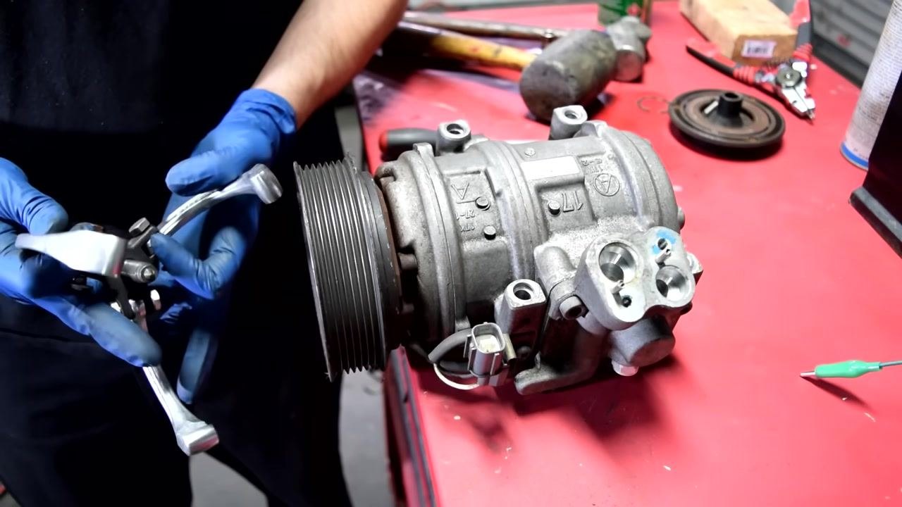 A person in blue gloves holds a two or three jaw puller near the AC compressor pulley, preparing to use it for pulley removal.