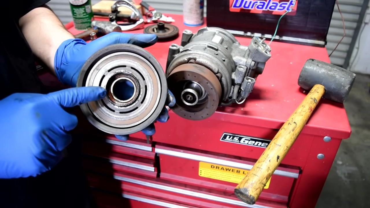 A person in blue gloves holds the removed AC compressor pulley, showing the bearing inside. The compressor and a rubber mallet are on the red tool chest.