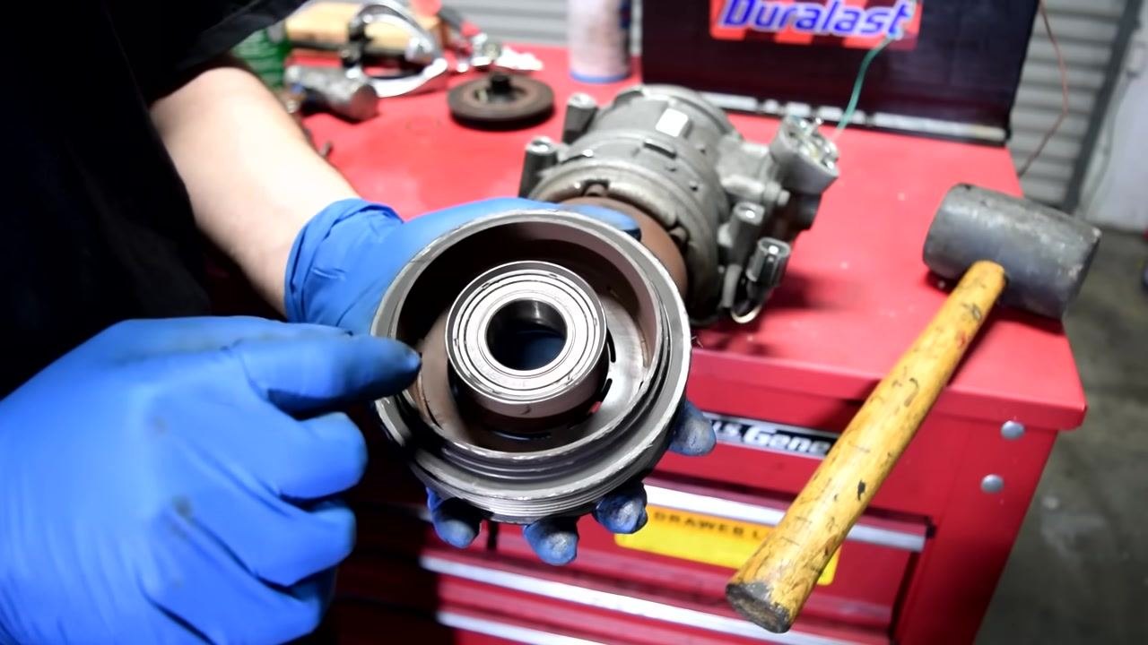 A gloved hand points to the outer lip of a bearing inside a pulley assembly, with a rubber mallet and AC compressor visible in the background.