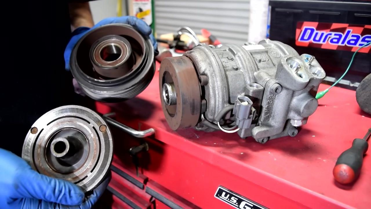 A gloved hand points to the stator/coil mounted on the AC compressor, with visible wiring and connectors.