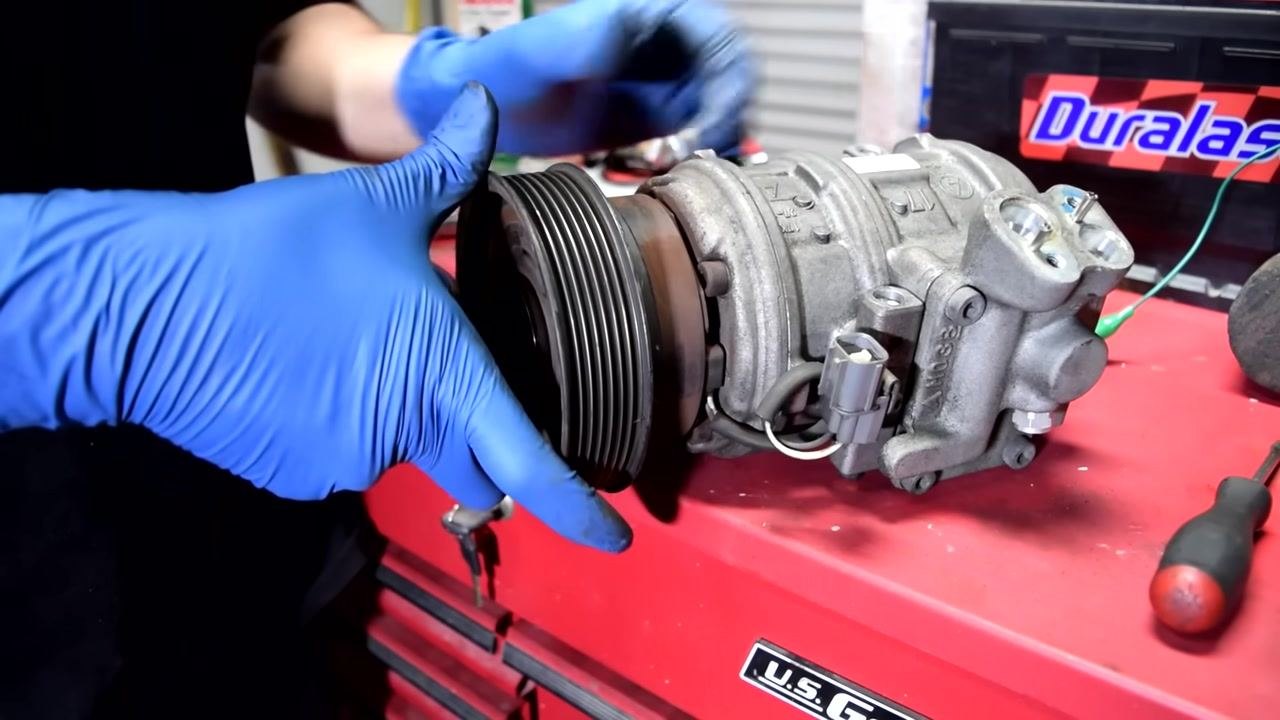 A gloved hand begins to fit the pulley onto the AC compressor shaft, showing the initial alignment.