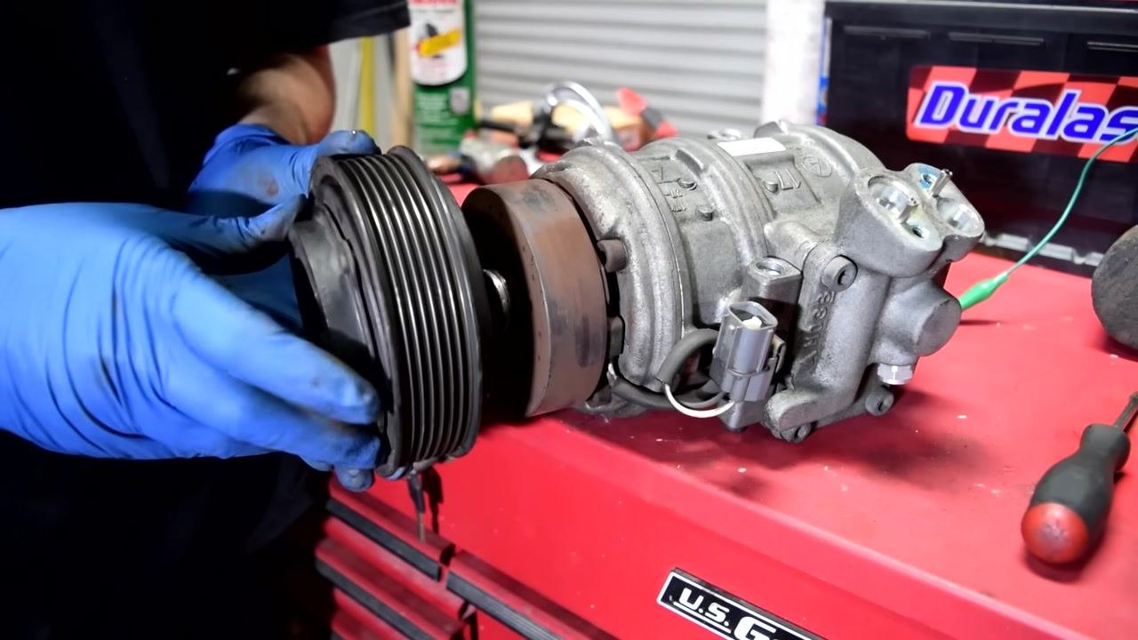 A gloved hand holds the pulley assembly on the AC compressor, preparing for coil removal, with the connector and wiring visible.