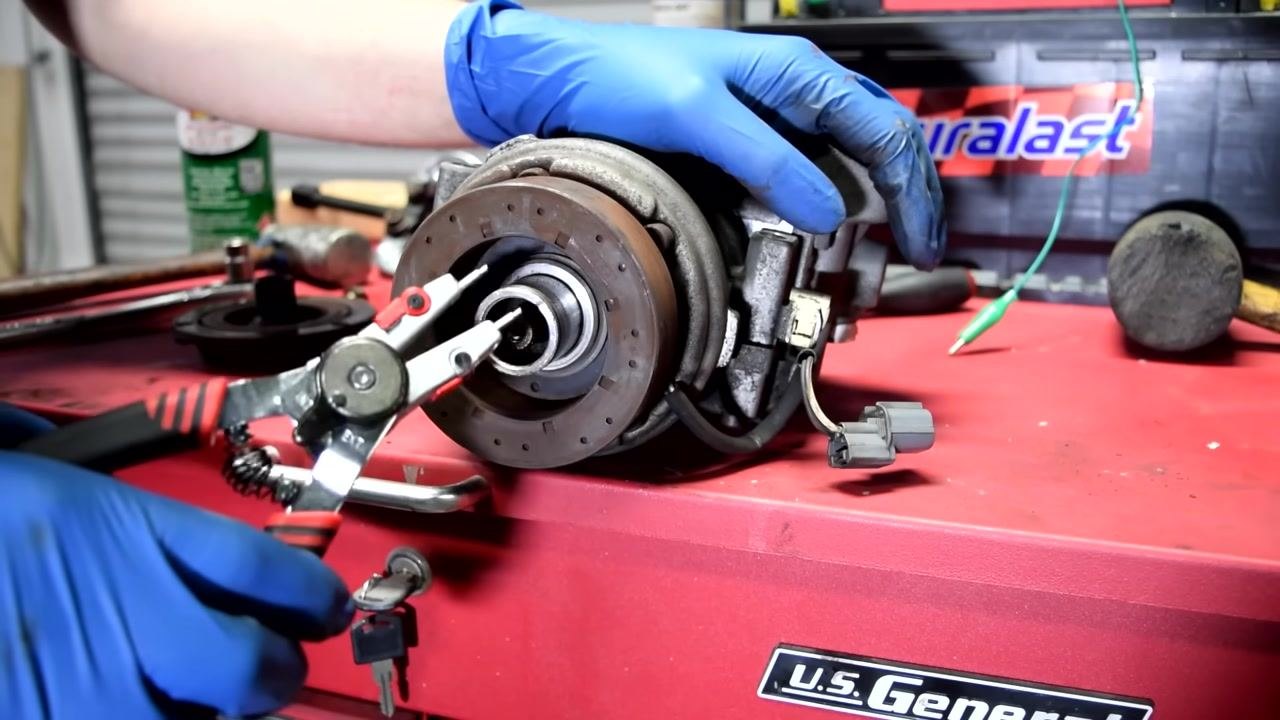 A gloved hand uses snap ring pliers to remove the retaining ring from the AC compressor shaft, with the compressor secured on a red tool chest.