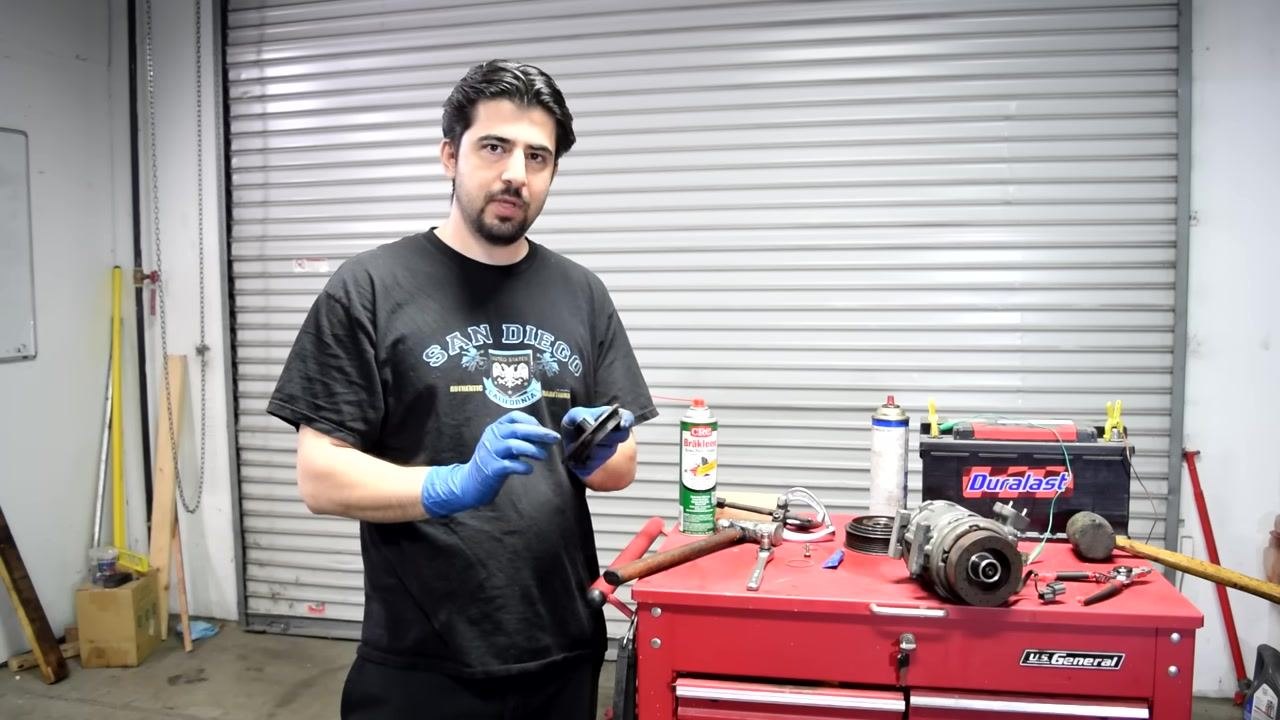 A gloved hand holds the clutch plate and the mounting bolt, preparing for installation. The AC compressor is visible in the background.
