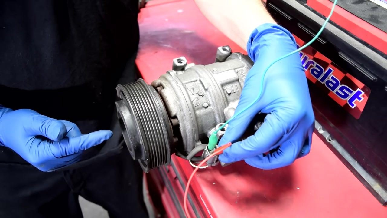 Close-up of the AC compressor's electrical connector, showing a single white wire and a gloved finger pointing to it.