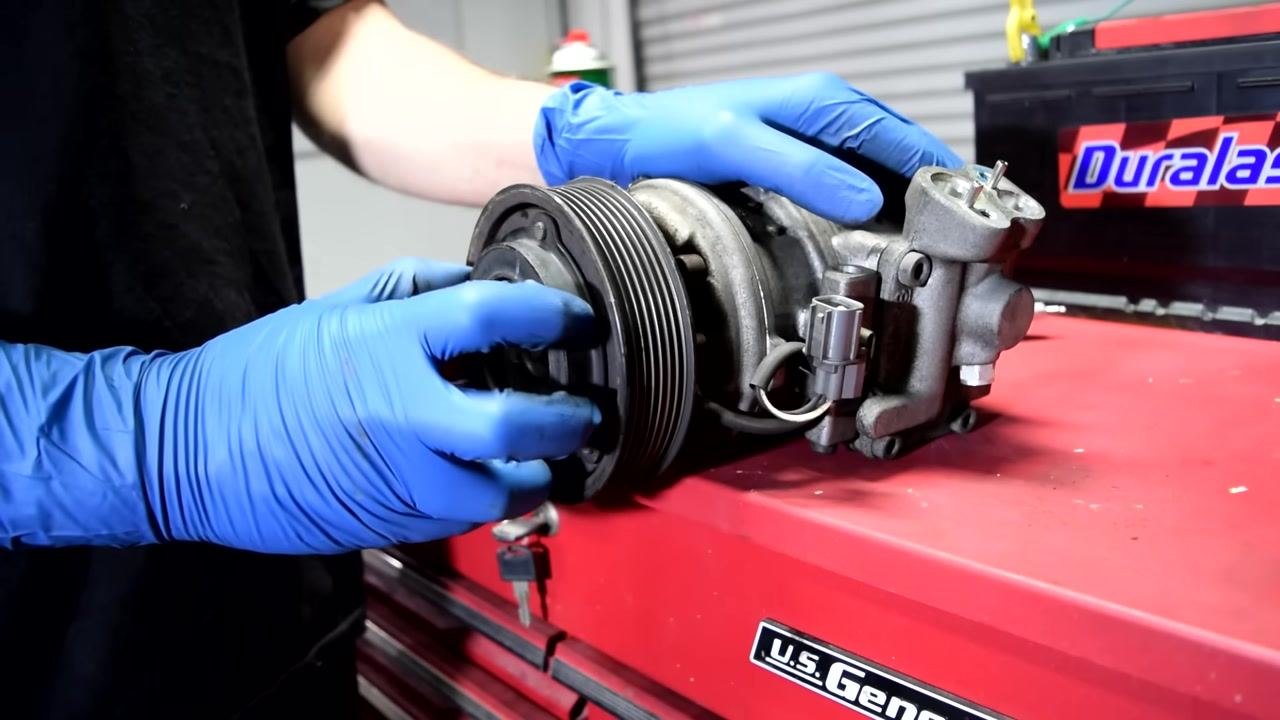 A person wearing blue gloves connects green and red alligator clips to the AC compressor's terminals, preparing to test the clutch coil.