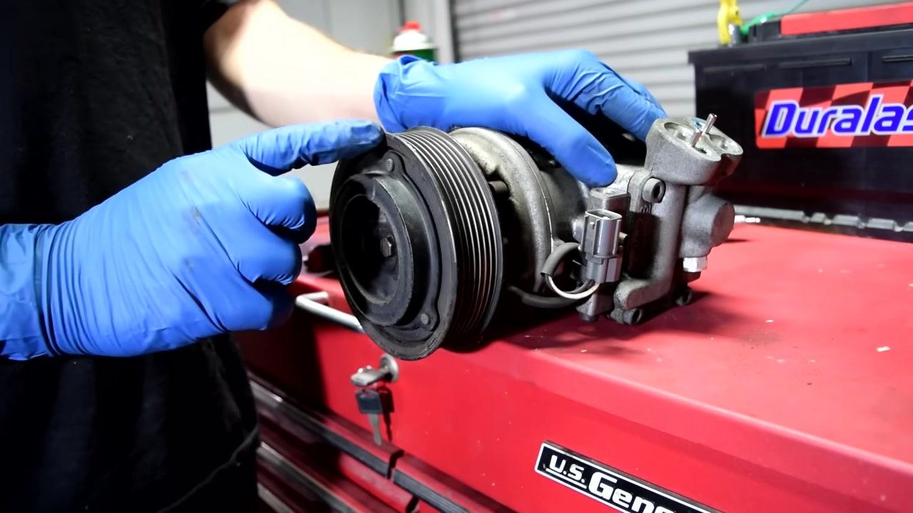 A person in blue gloves holds the AC compressor, pointing to the pulley and bearing area where you should listen for issues.