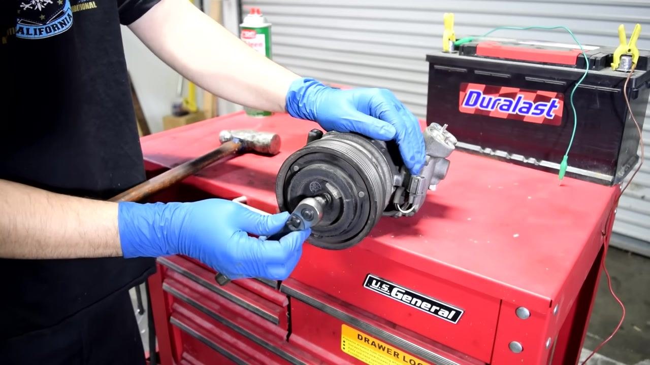 A person wearing blue gloves uses a ratchet and socket to loosen the central bolt on the AC compressor clutch. A hammer is nearby on the red tool chest, and a Duralast battery is in the background.
