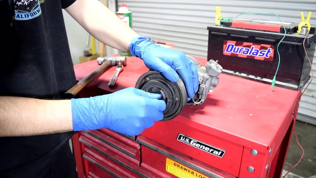 A person in blue gloves holds the removed AC compressor clutch plate in one hand, with the compressor and pulley visible on the workbench.