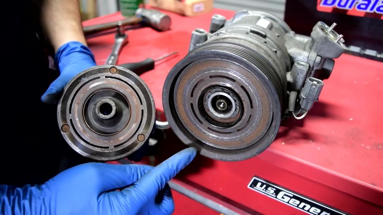 A person wearing blue gloves points to the contact surface on the AC compressor pulley and the compressor itself. The pulley is held in one hand, and the compressor is on a red tool chest.