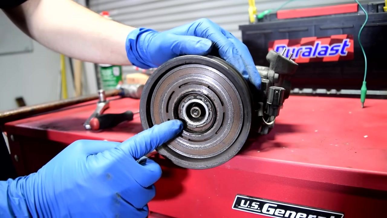 A person in blue gloves points to the snap ring on the AC compressor pulley, preparing to remove it with snap ring pliers.