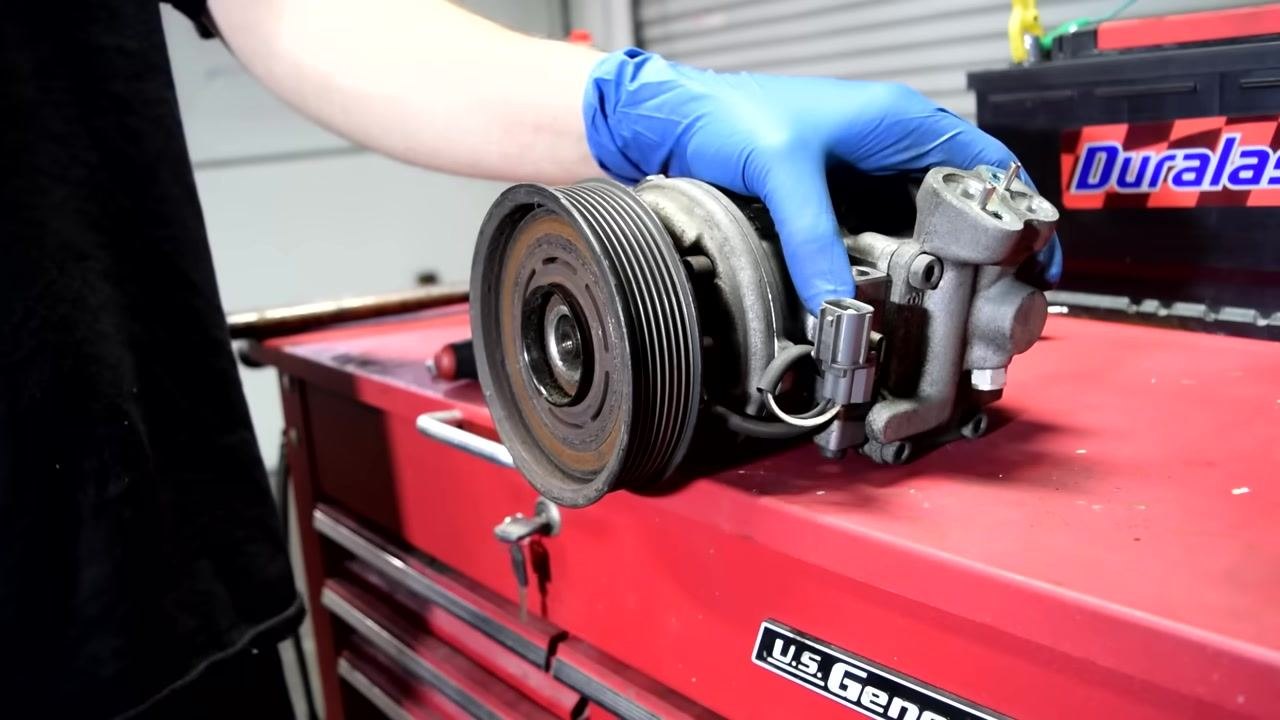 A person in blue gloves attempts to pull the pulley off the AC compressor by hand. The pulley is still attached to the shaft.
