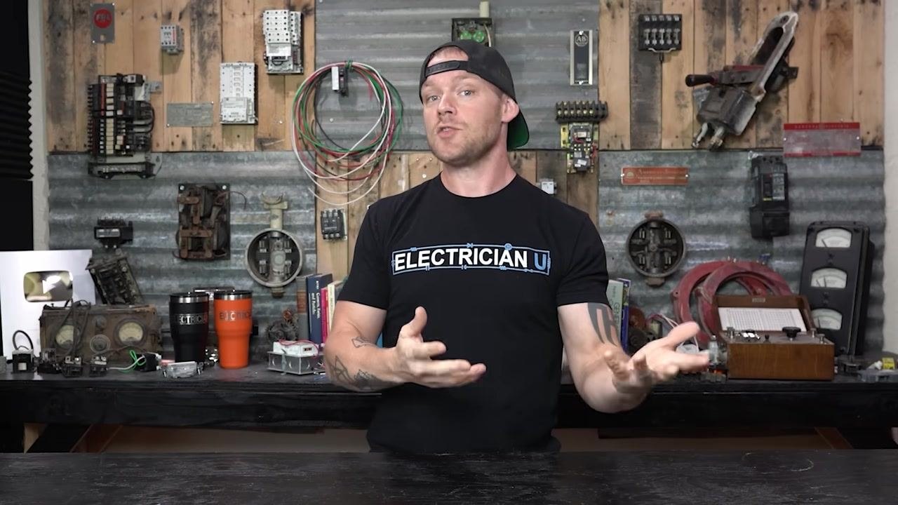 Presenter in workshop discussing breaker overheating, surrounded by electrical panels, breakers, and tools