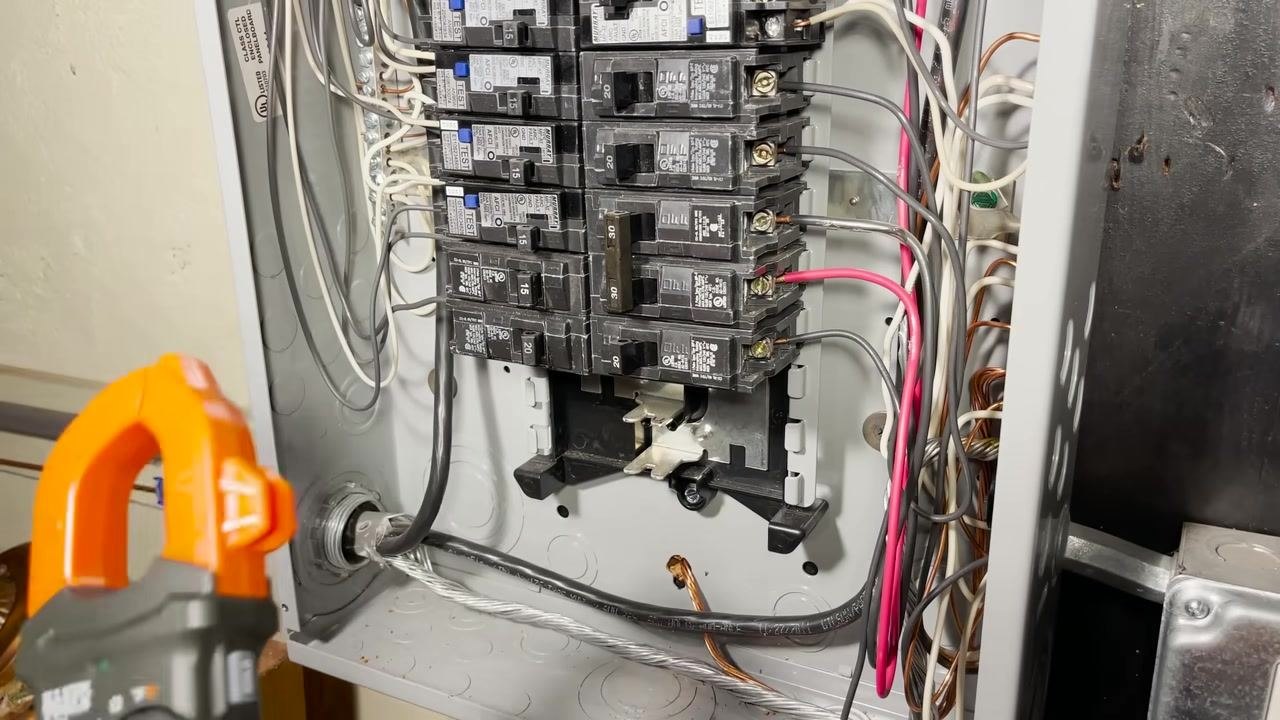 Open electrical panel with multiple breakers, visible wiring, and a clamp meter being used for voltage testing
