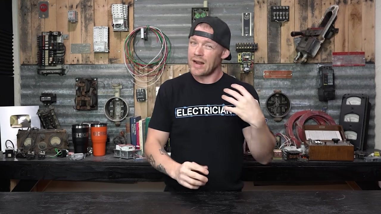 Presenter in workshop, explaining breaker overheating causes, replacement options for legacy panels, and specialty breakers. Various electrical components, replacement breakers, and tools are visible on the workbench and wall.
