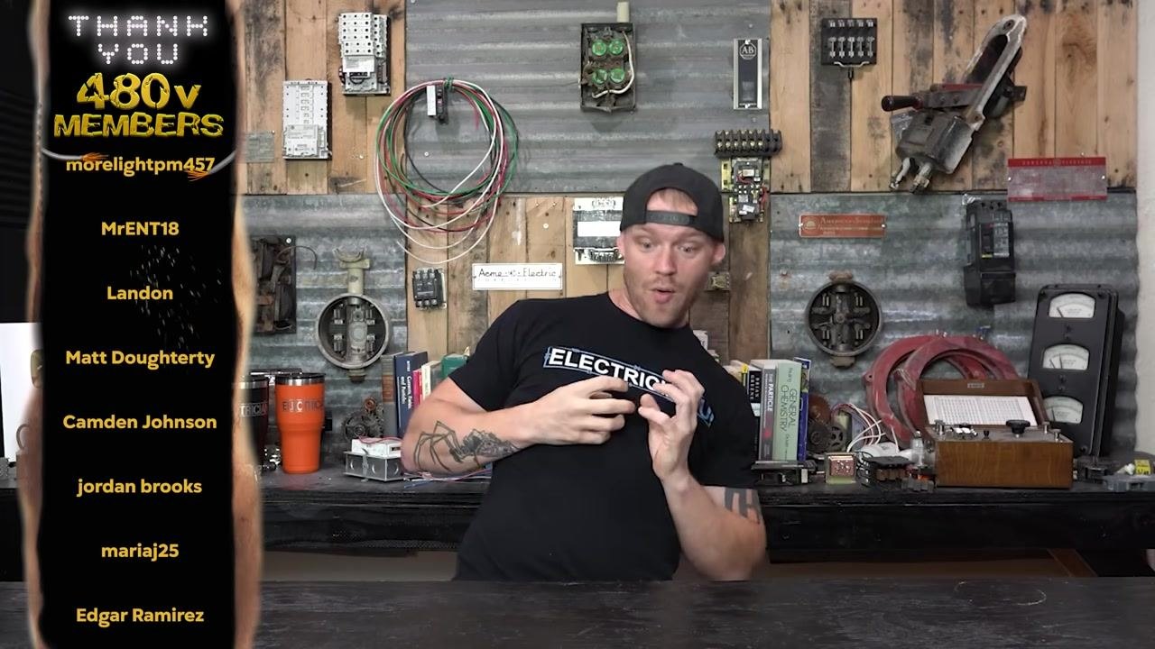 A person in a workshop setting, standing behind a workbench, wearing a black "ELECTRICIAN U" shirt and a backwards cap. The background features various electrical components, books, and tools mounted on a wall. The person is gesturing with their hands, emphasizing a point.
