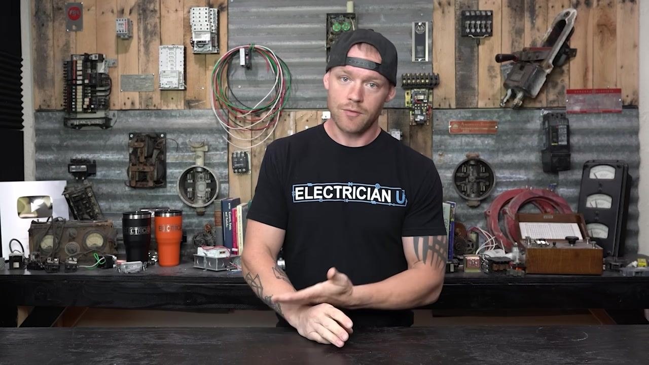 A person in a workshop, wearing an "ELECTRICIAN U" shirt, explaining breaker testing steps and mechanical failure symptoms. Various electrical components and tools are visible in the background.