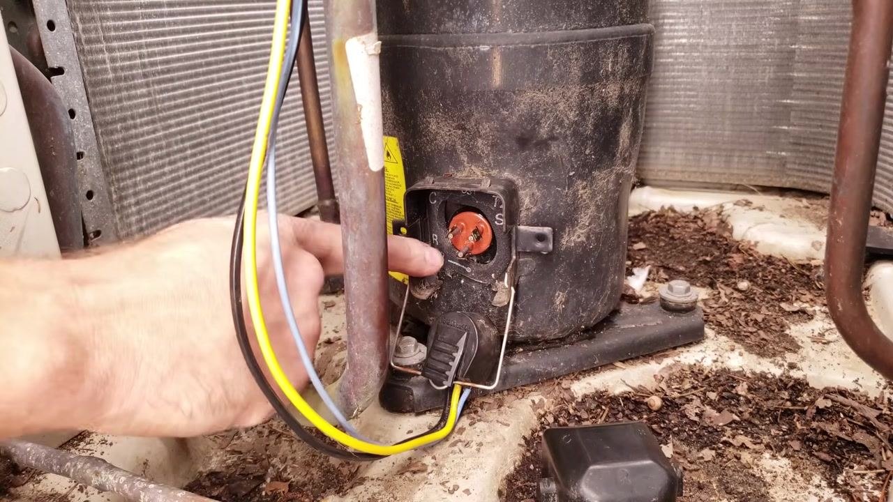 Technician pointing at the exposed compressor plug terminals with yellow and blue wires visible