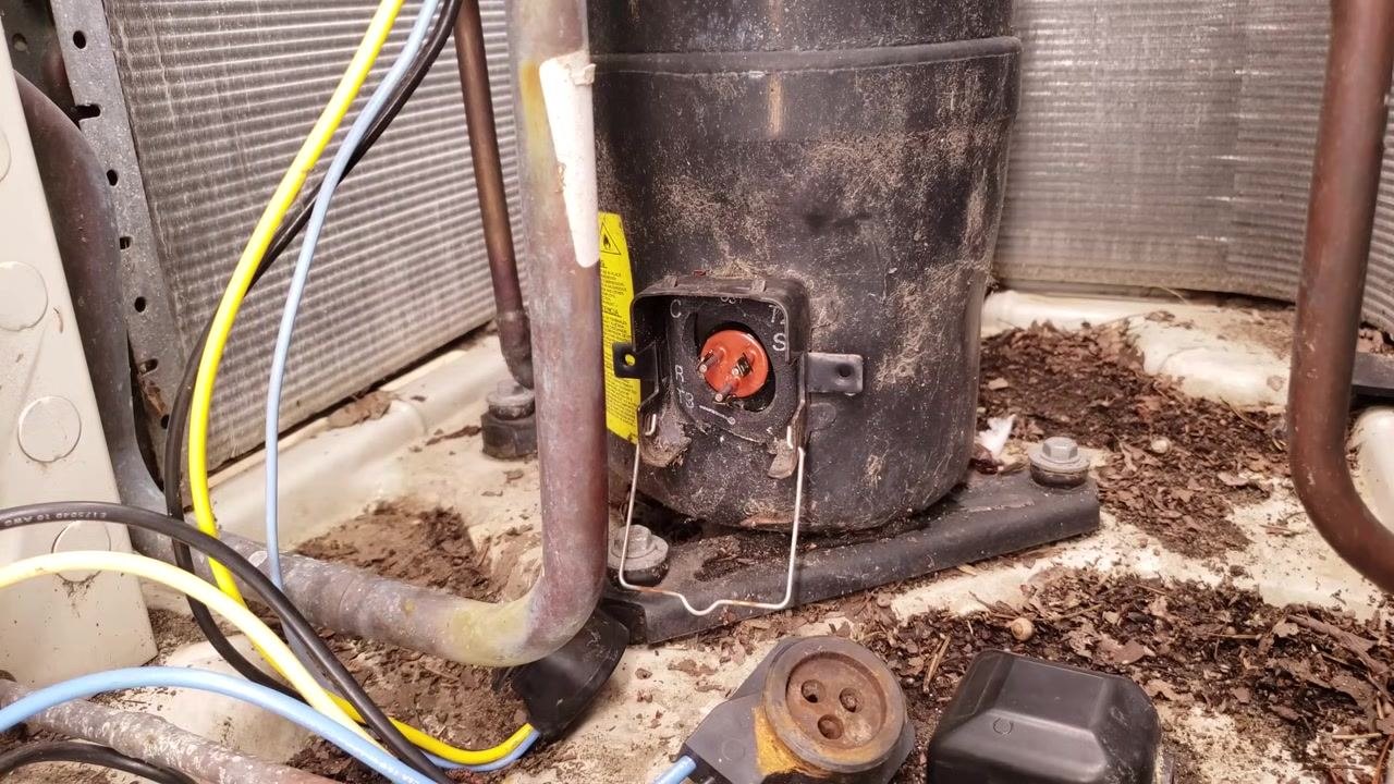 Exposed compressor terminals labeled C, R, S with the removed plug and cover on the unit base, yellow and blue wires, copper tubing, and mounting bolts visible