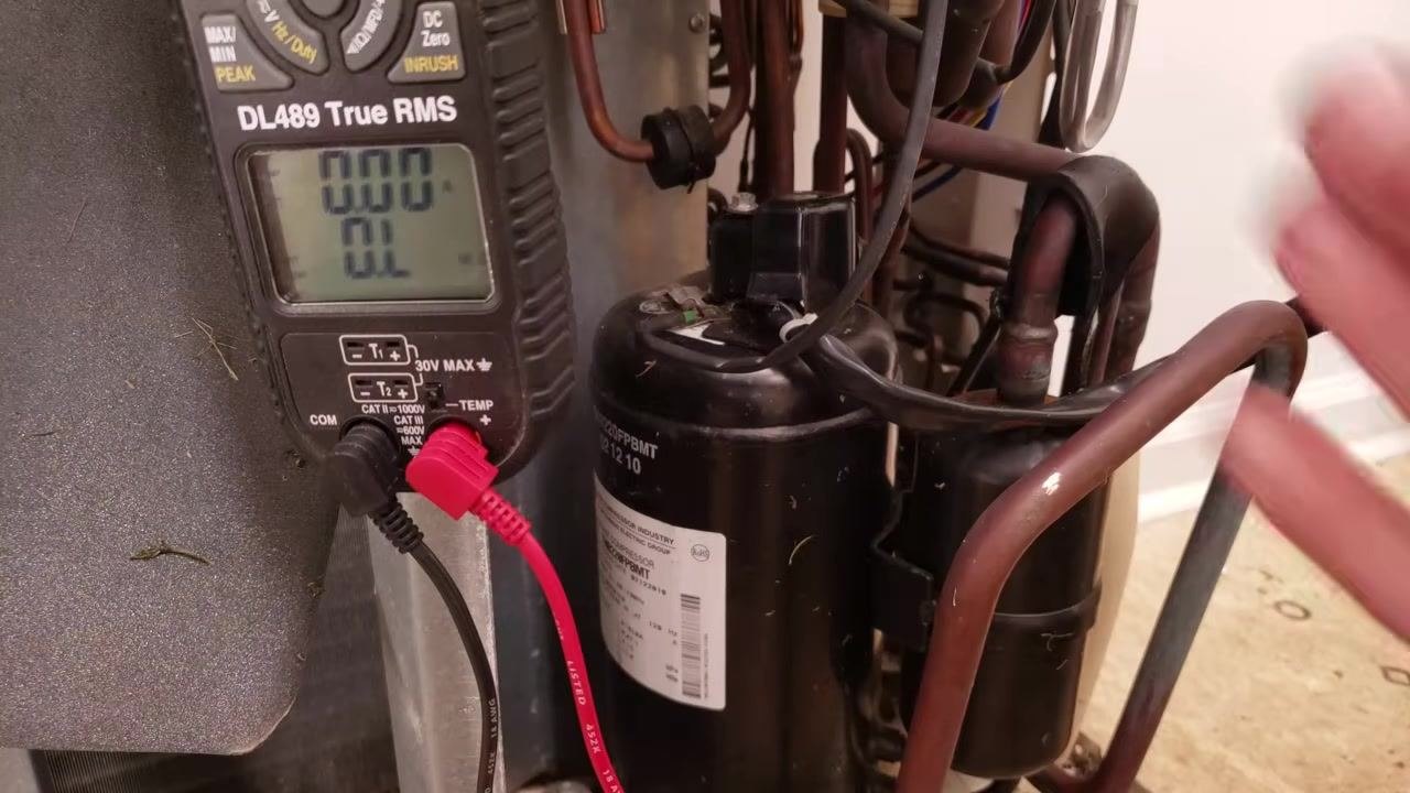 Mini-split compressor with a multimeter connected showing "OL" on the display, copper tubing, wires, and compressor label visible