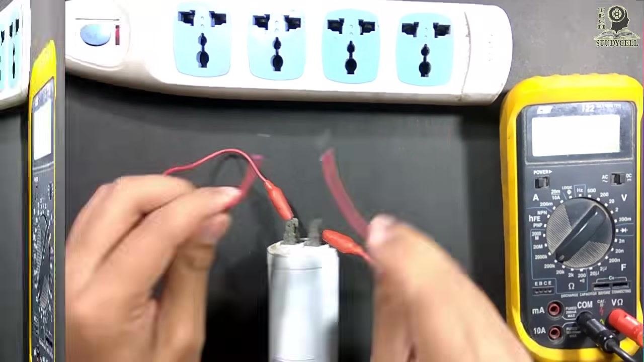 A start capacitor with two red wires attached to its terminals using alligator clips, with hands holding the wires above the capacitor and a power strip and multimeter visible on the table