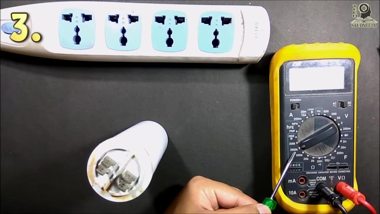 A digital multimeter with its dial set to the 200kΩ resistance range, red lead in the VΩ port, black lead in the COM port, with a screwdriver pointing at the dial and a start capacitor and power strip visible
