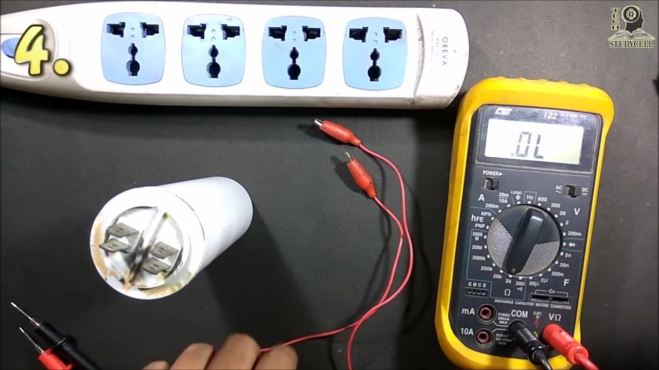A large white capacitor, a yellow digital multimeter displaying "OL," red test leads with alligator clips, and a power strip arranged on a black work surface, with the dial set to the capacitance (F) range