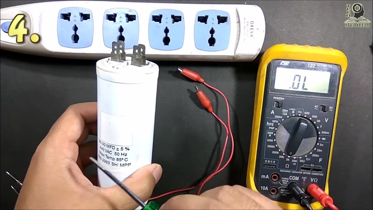 A hand holding a large white capacitor while another hand touches a screwdriver to the terminals to discharge it, with the multimeter set to "OL" and red test leads with alligator clips visible
