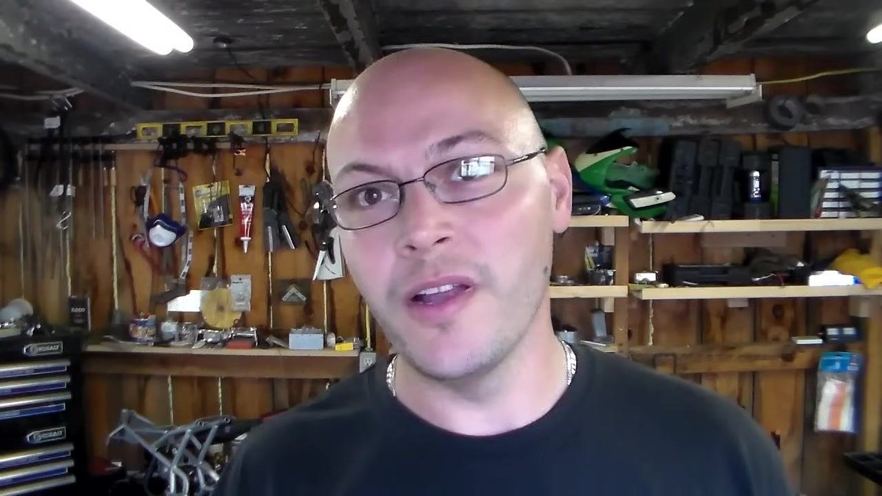 Person in a workshop, speaking directly to the camera with tools and shelves in the background.