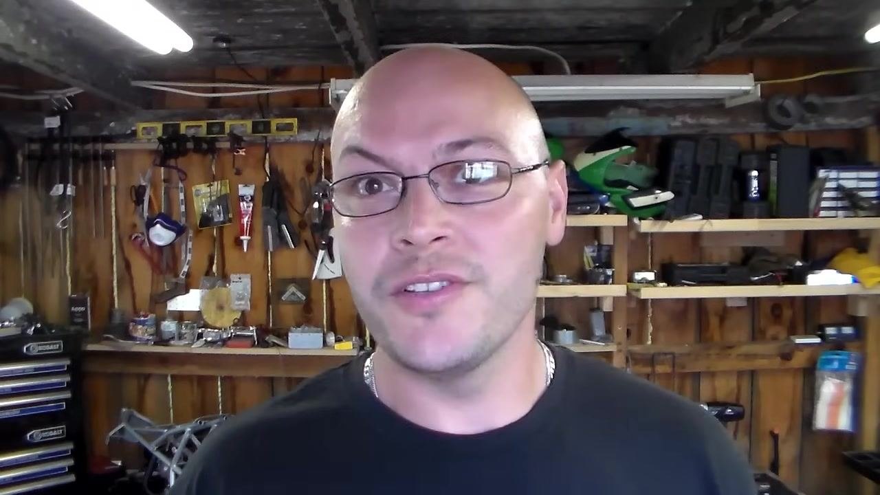 Presenter in a workshop, discussing replacement options for relays and capacitors. Shelves with tools, helmets, and supplies in the background.
