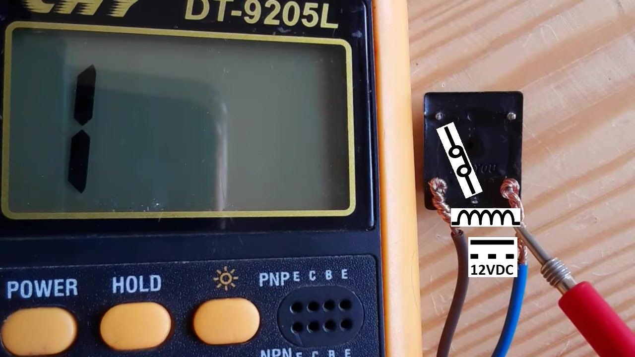 Relay on a wooden surface with two wires connected to the coil terminals, a "12VDC" label, and a closed switch schematic symbol overlaid