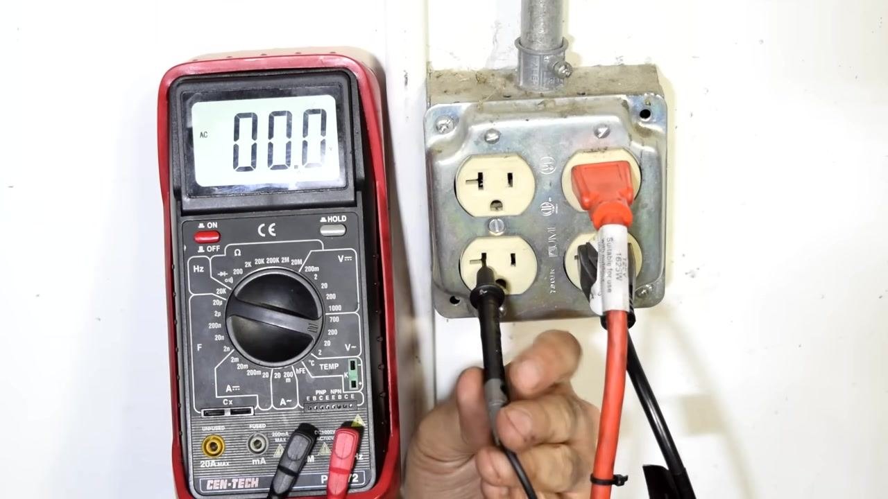 Close-up of a wall socket with the black test lead in the neutral slot and the red test lead about to be inserted into the hot slot; the multimeter displays 0.00 AC volts.