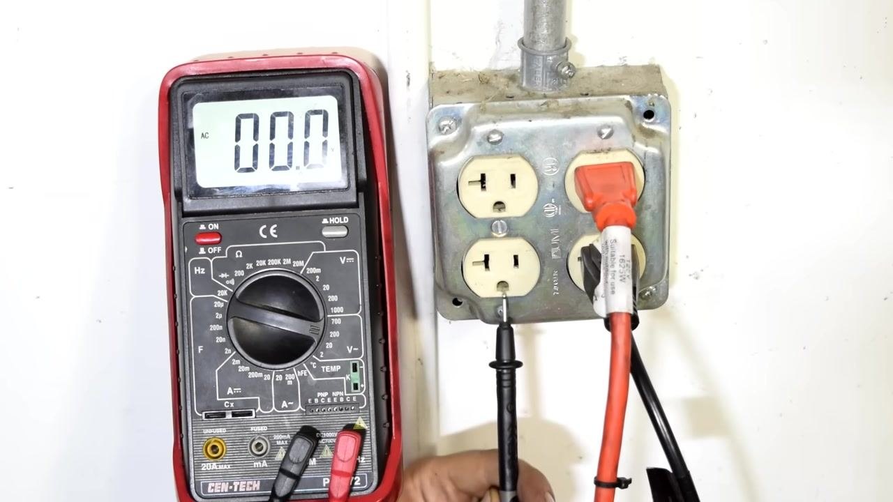 Close-up of a wall socket with the black test lead inserted into the ground slot; the multimeter displays 0.00 AC volts.