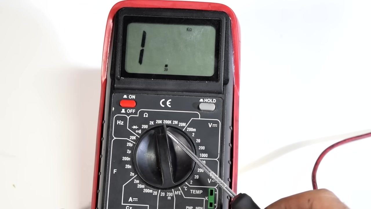 A multimeter display shows 9.87, indicating the measured resistance in kilo-ohms (kΩ), with the test leads connected to a spark plug wire and both hands holding the wire and probe.