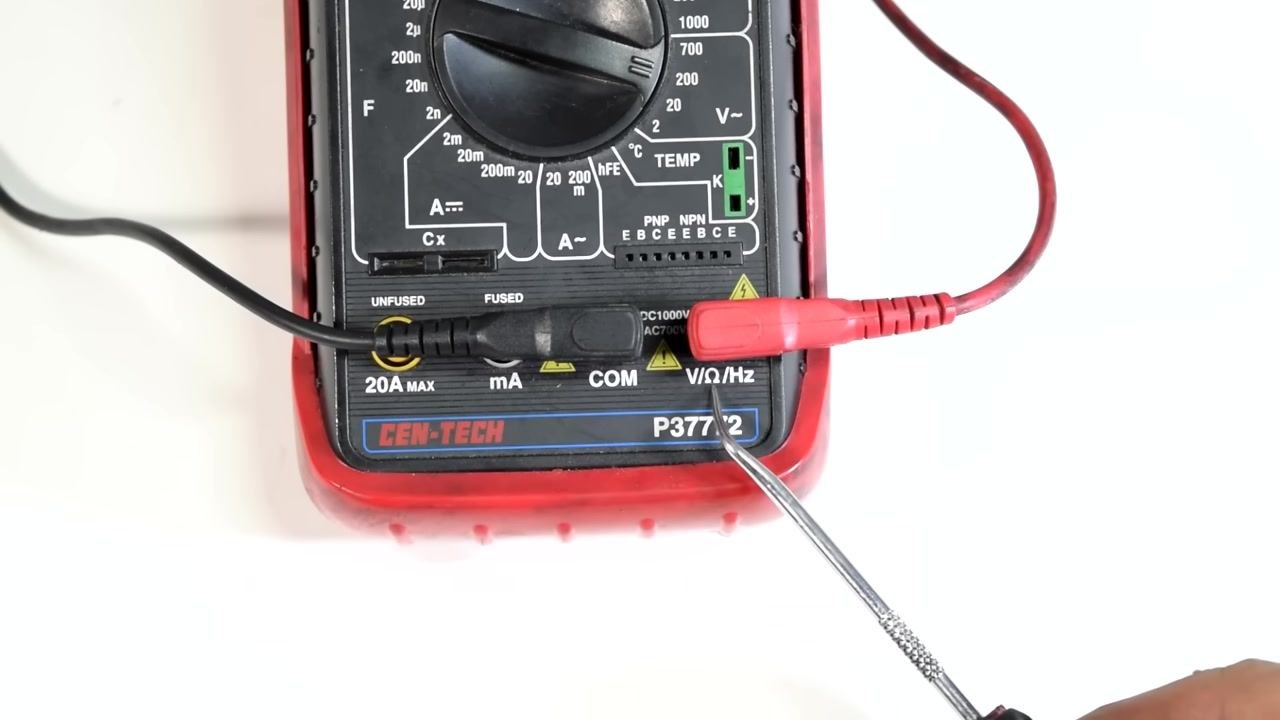 A multimeter with test leads connected to a spark plug wire using alligator clips; the display shows "1", indicating an open circuit or out-of-range measurement, with both hands holding the wire.