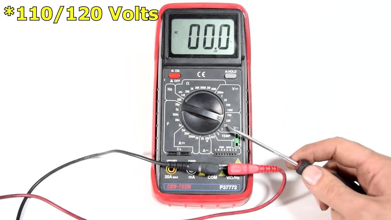 A digital multimeter set to the 200V AC range, with a hand holding a metal pointer and the display reading 0.00. Large yellow text reads "*110/120 Volts".