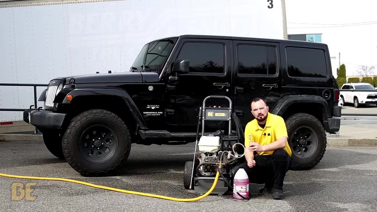 Person crouched next to a BE Power Washer, holding the soap injector line and inspecting the screen at the end, with a black Jeep and cleaning solution nearby