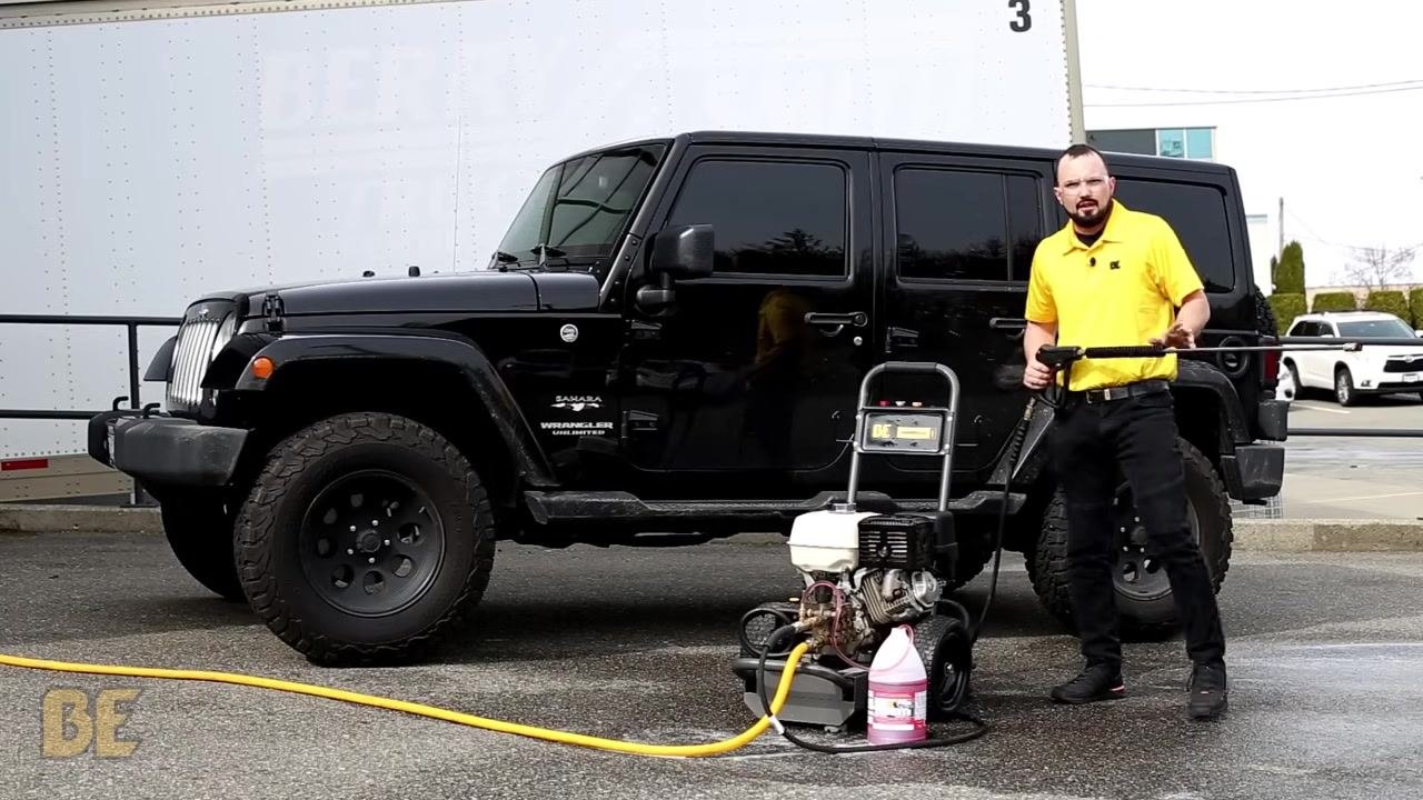 Person in a yellow shirt standing next to a black Jeep Wrangler and a BE Power Washer, preparing to rinse the vehicle, with soap solution and equipment visible in the foreground