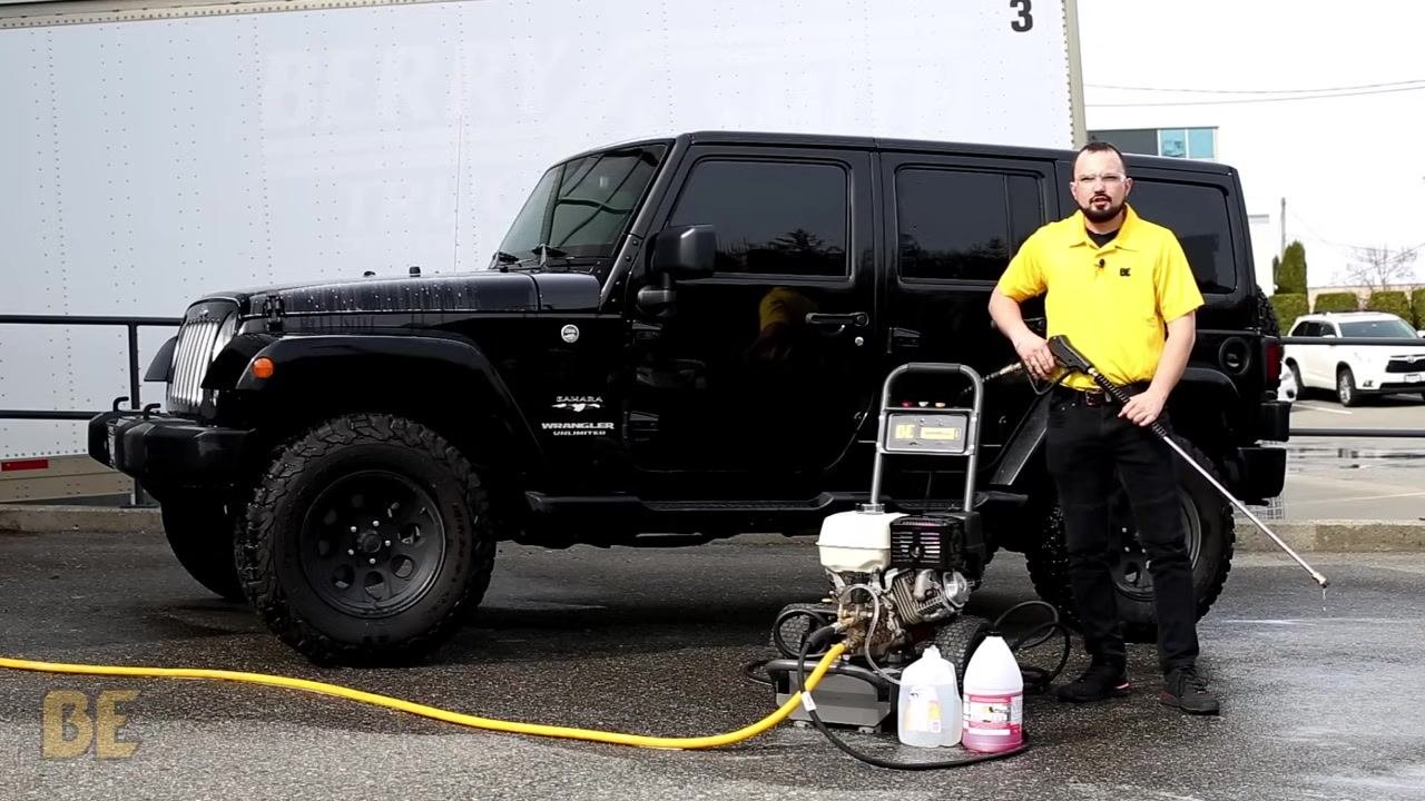 Person in a yellow shirt holding the spray wand next to the BE Power Washer and black Jeep Wrangler, with the soap injector hose in a clean bucket of water during the flushing process