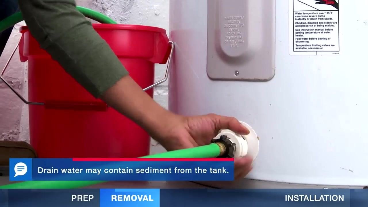 A person kneeling next to a water heater, attaching a green garden hose to the drain valve, with a red bucket nearby and a warning label visible on the heater.