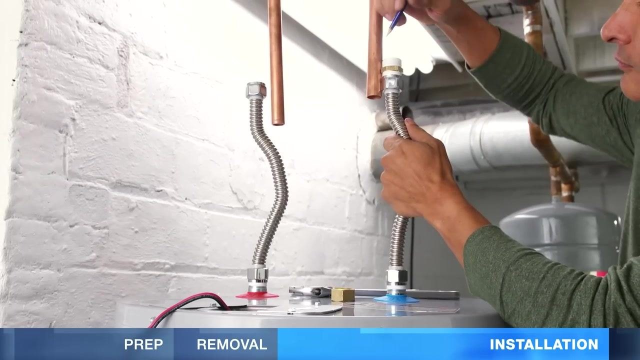 A person holding a flexible hose up to a copper water supply pipe above the new water heater, marking the pipe for cutting, with both hot and cold lines visible.