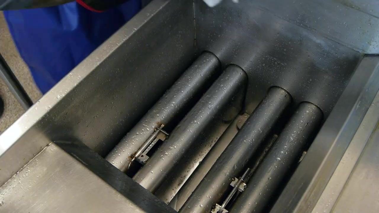 Person wearing black protective gloves using a rag and a metal poker to wipe between the heating flumes inside a commercial fryer basin, with all surfaces visibly clean and wet.