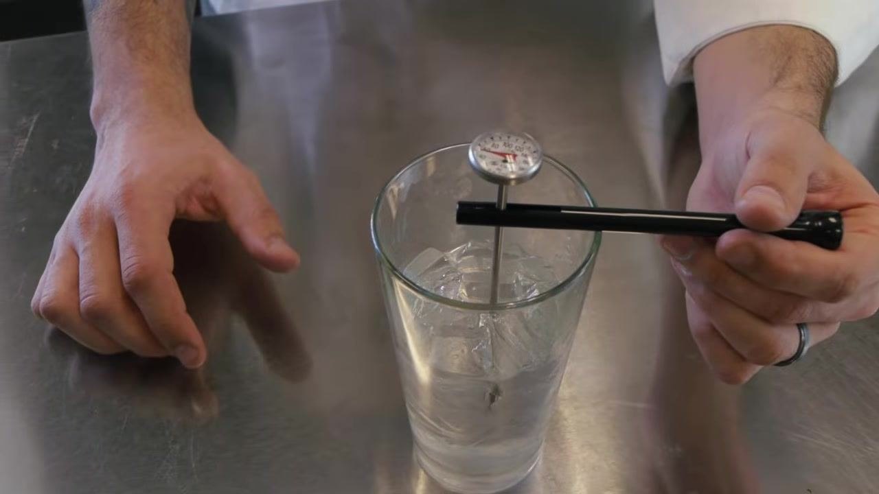 Dial probe thermometer suspended in a glass of ice water, needle visible, reading above 30°F