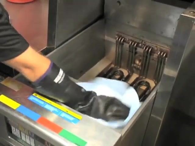 A gloved hand wipes the inside of the fryer vat with a blue towel.