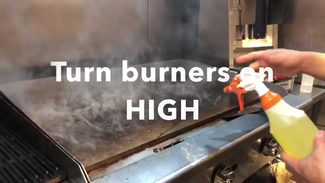A hot, steaming flat-top grill with a person holding a spray bottle containing yellow liquid. Large on-screen text reads "Turn burners on HIGH."