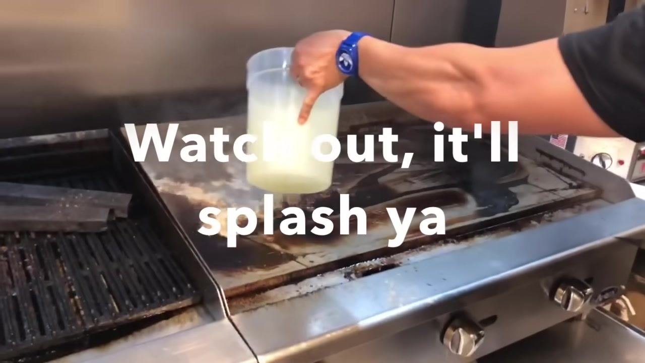 A person pouring a pale yellow lemon water solution from a large container onto the hot grill. On-screen text reads "Watch out, it'll splash ya."
