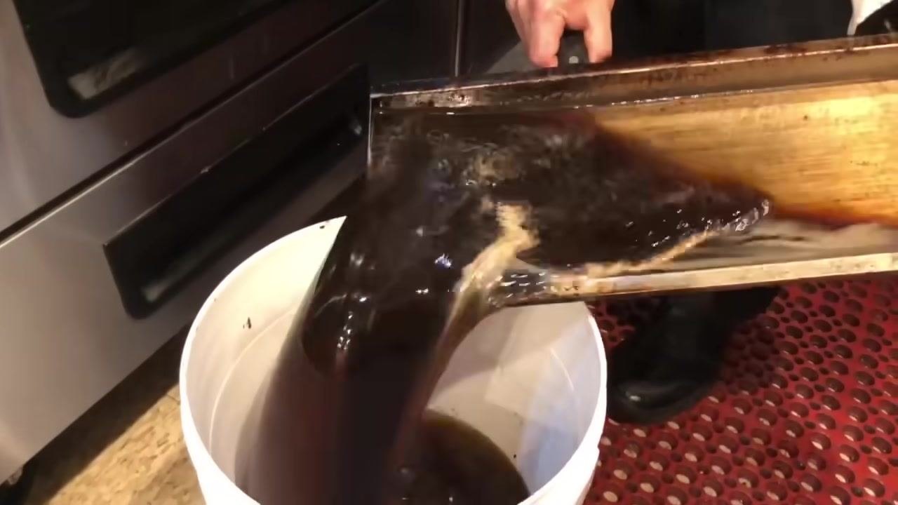 A person pouring dark, dirty water from the grill's waste tray into a white bucket.